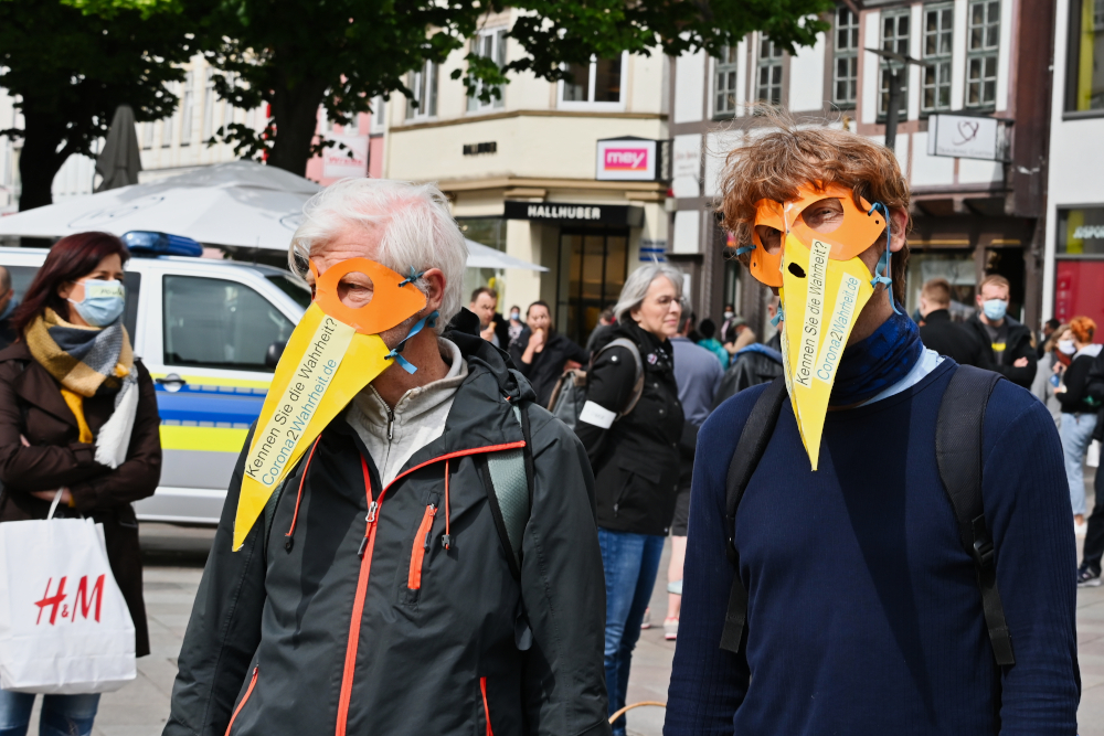 Auf dem Bild sind zwei Männer mit selbst gebastelten Krähen-Masken zu sehen. Auf den Masken steht "Kennen Sie die Wahrheit? Corona2Wahrheit.de". Im Hintergrund ist die Göttinger Innenstadt, weitere Demonstrant:innen und Passant:innen.
